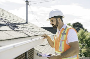 Why Was My Roof Damage Claim Not Approved By Insurance? | Pride Roofing LLC. Image of a male roof inspector in a hard hat, holding a clipboard, standing on the steps of an old rundown house, checking for the state of the roof.