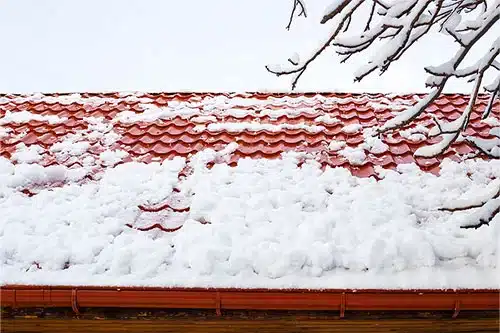 How Snow and Ice Impact Your Roof | Pride Roofing LLC. Image of a red metal-tiled roof after heavy snowfall in winter.